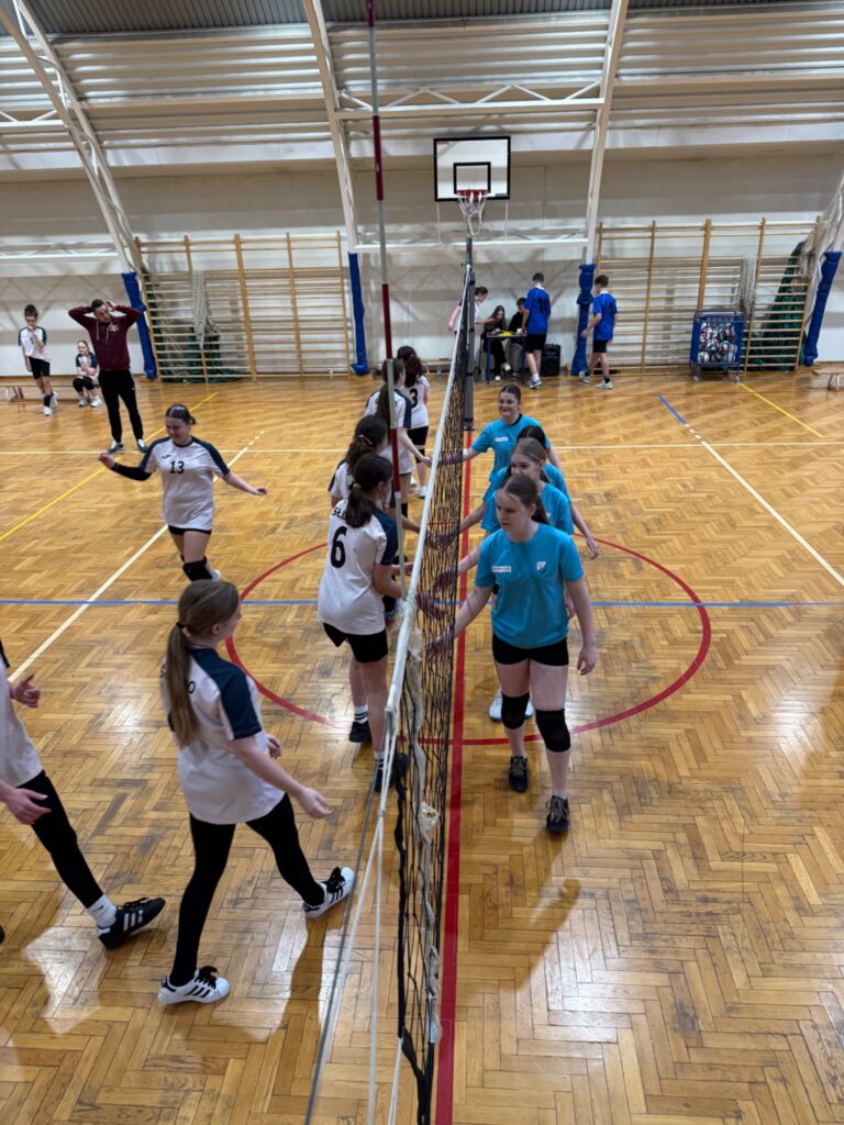 Drużyny siatkarskie w białych i turkusowych koszulkach ściskają dłonie przy siatce po zakończeniu finałów w hali.