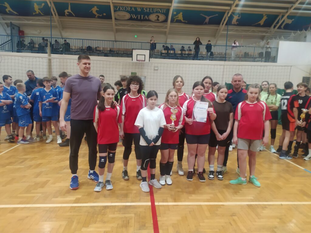 Grupa młodych siatkarzy i siatkarek w czerwonych koszulkach stoi na boisku podczas finałów siatkówki, trzymając trofeum i dyplom.