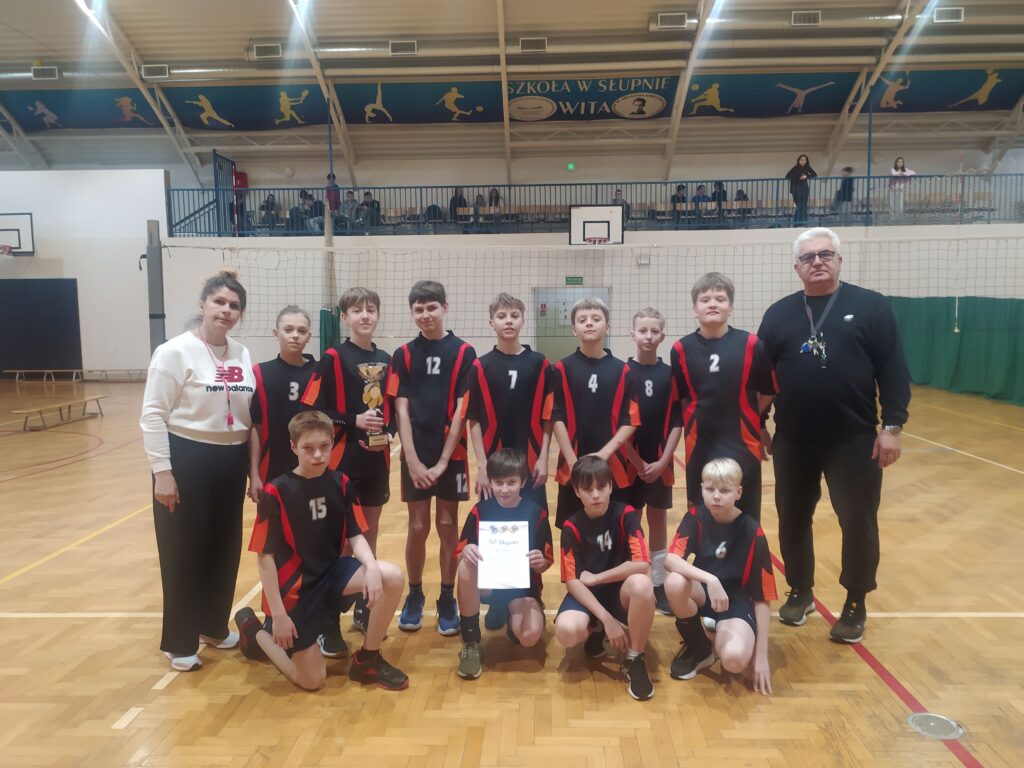 Grupa młodych siatkarzy w czarnych i czerwonych strojach z numerami stoi na boisku w hali wraz z trenerami, trzymając puchar i dyplom po finałach siatkówki.