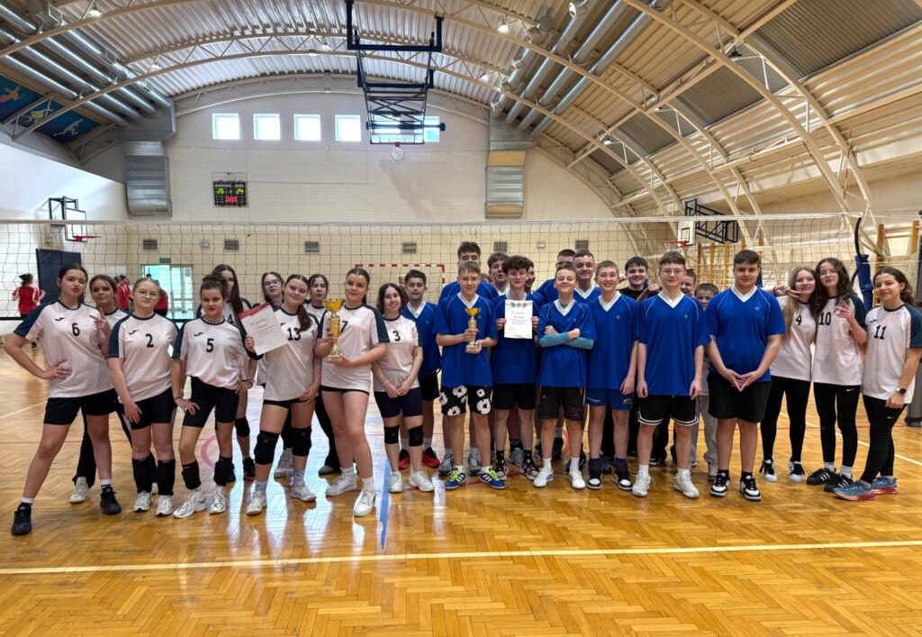 Grupa młodych siatkarzy i siatkarek stoi na boisku w hali podczas finałów siatkówki, trzymając puchar i dyplomy.