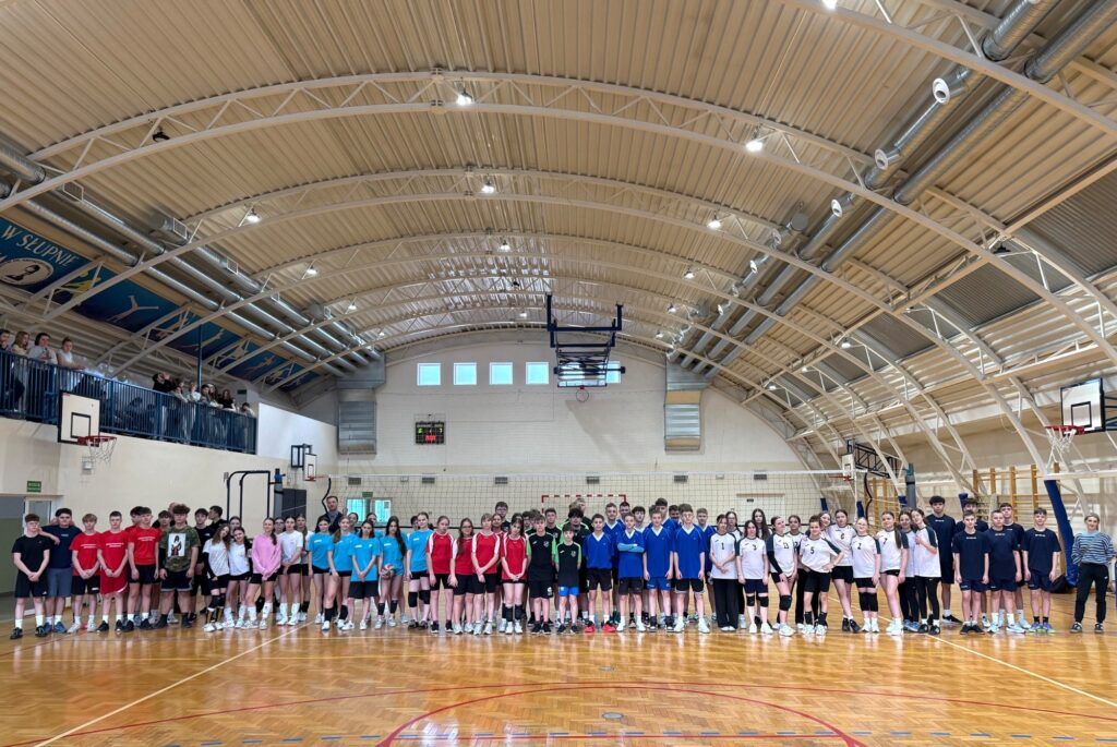 Duża grupa młodych zawodników w kolorowych koszulkach stoi w rzędzie na boisku hali podczas finałów siatkówki, z widzami na trybunach po lewej.