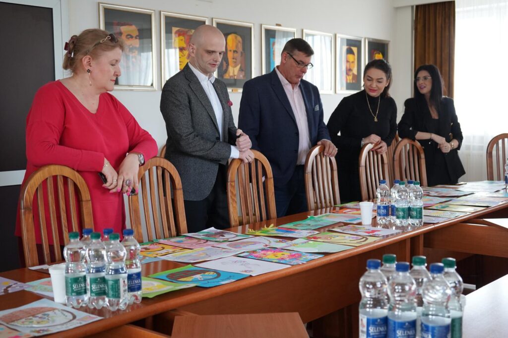 Kilka osób stojących w sali konferencyjnej za długim stołem, na którym rozłożone są kolorowe kartki wielkanocne, podczas rozstrzygnięcia konkursu Powiatu Płockiego.