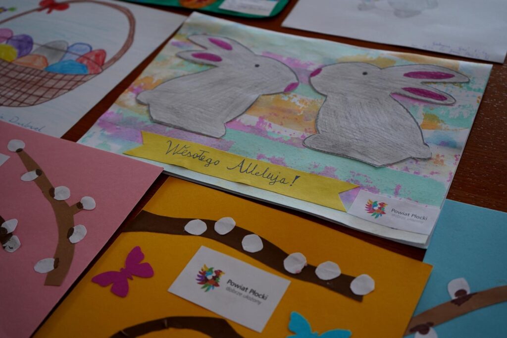 Kolorowe kartki wielkanocne rozłożone na stole, w centrum karta z dwoma szarymi zajączkami na pastelowym tle i żółtą wstęgą z napisem „Wesołego Alleluja!”, z widocznym logo Powiatu Płockiego.