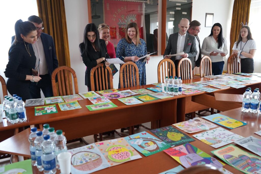 Grupa osób ogląda kartki wielkanocne rozłożone na stole podczas rozstrzygnięcia konkursu Powiatu Płockiego.