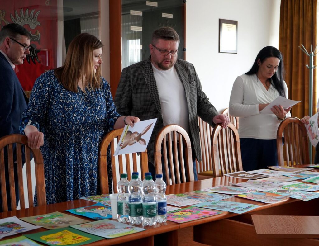 Grupa osób ogląda kolorowe kartki wielkanocne rozłożone na stole podczas rozstrzygnięcia konkursu Powiatu Płockiego.