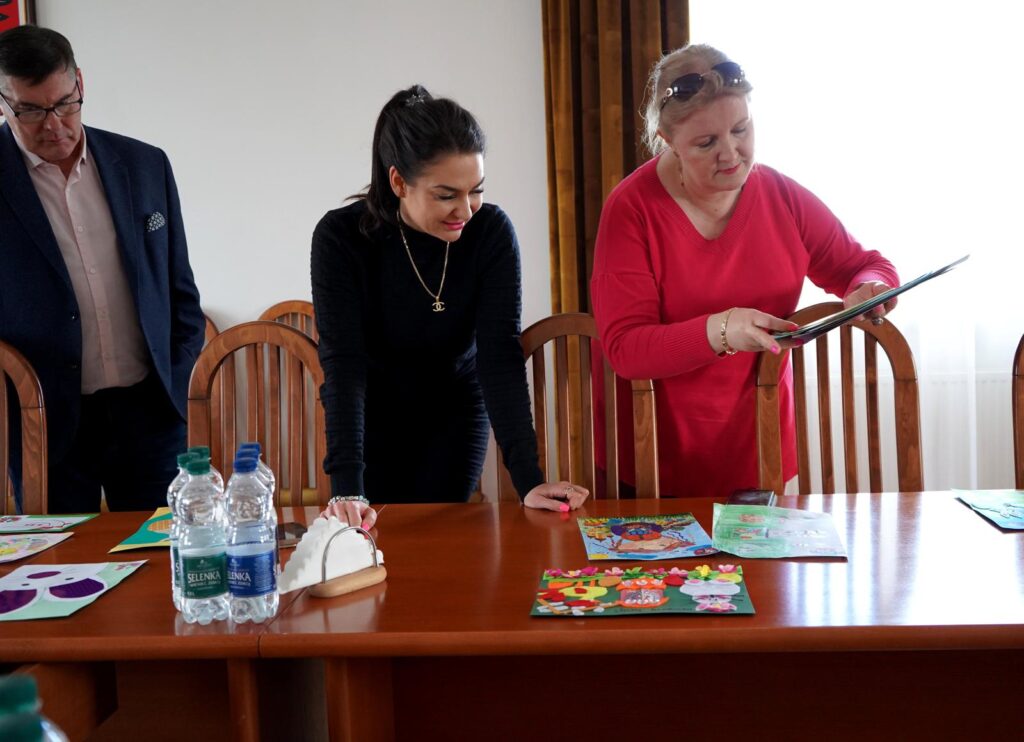 Trzy osoby stoją przy stole i oglądają kartki wielkanocne rozłożone na stole podczas rozstrzygnięcia konkursu Powiatu Płockiego.