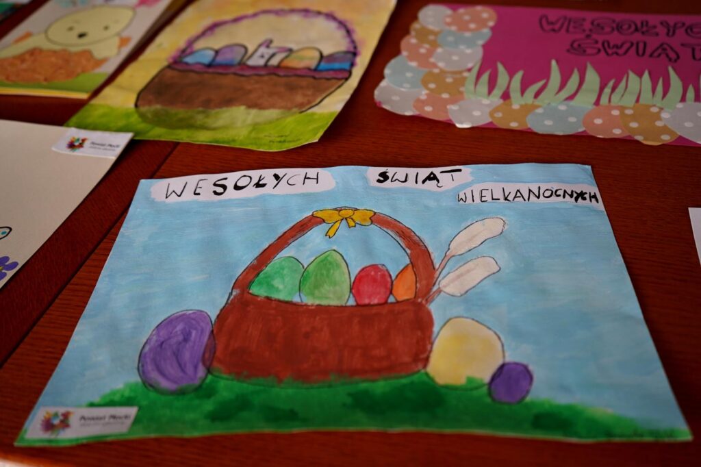 Kartka wielkanocna przedstawiająca brązowy koszyk wypełniony kolorowymi jajkami na zielonej trawie i błękitnym niebie, z napisem „Wesołych Świąt Wielkanocnych”.