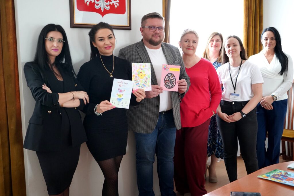 Grupa osób stoi w biurze i prezentuje kartki wielkanocne, związane z rozstrzygnięciem konkursu na kartkę wielkanocną Powiatu Płockiego.