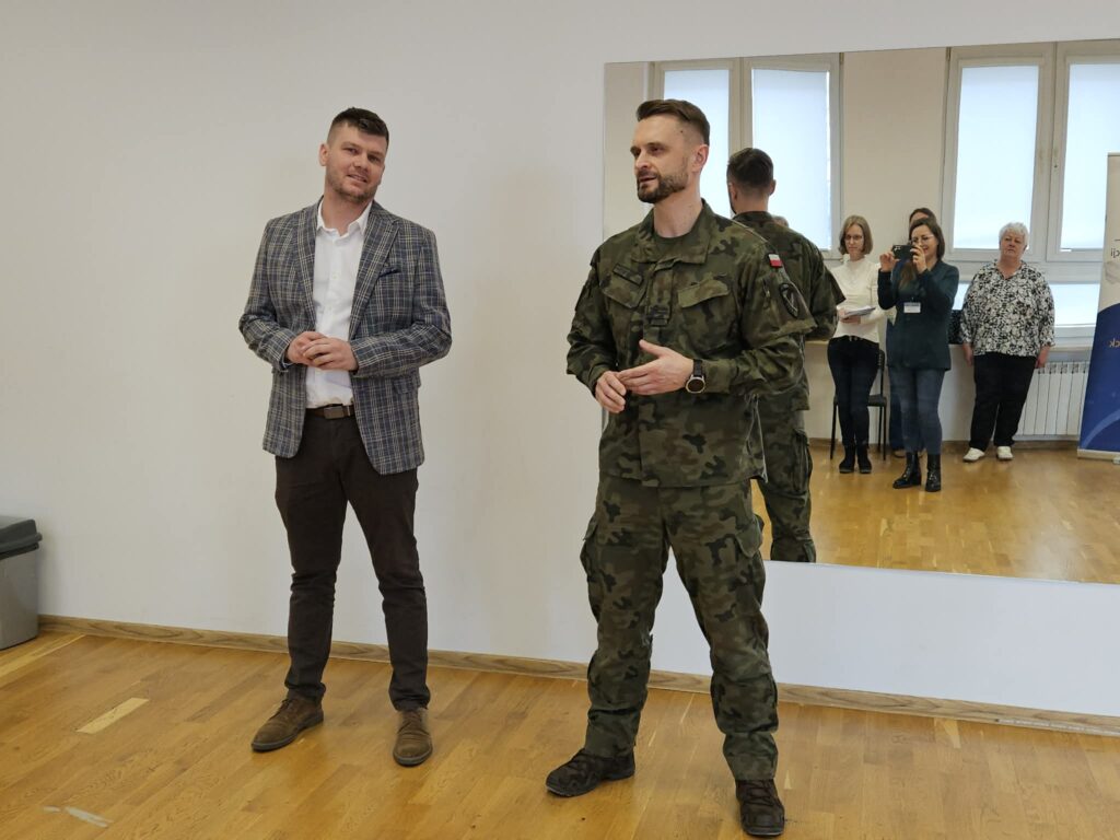 Dwóch mężczyzn stoi w sali podczas zakończenia kwalifikacji wojskowej w Powiecie Płockim: jeden w kratkowanej marynarce i białej koszuli, drugi w mundurze wojskowym, a w tle obserwatorzy.