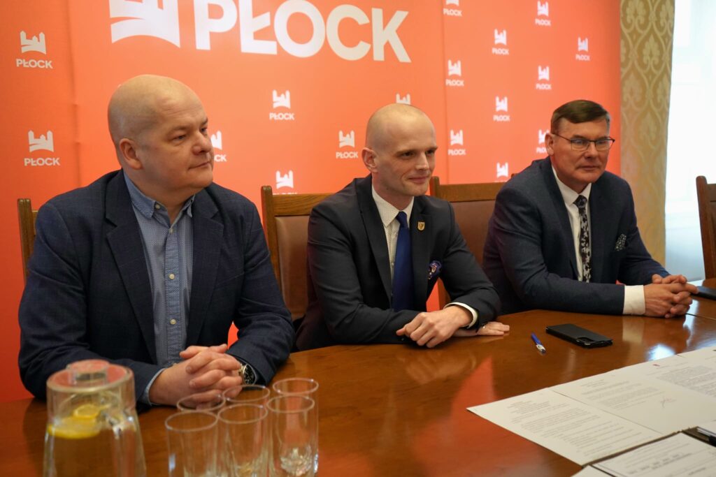 Trzech mężczyzn w garniturach siedzi przy stole w sali konferencyjnej, na tle pomarańczowego tła z logotypem Płocka i rozłożonymi dokumentami.