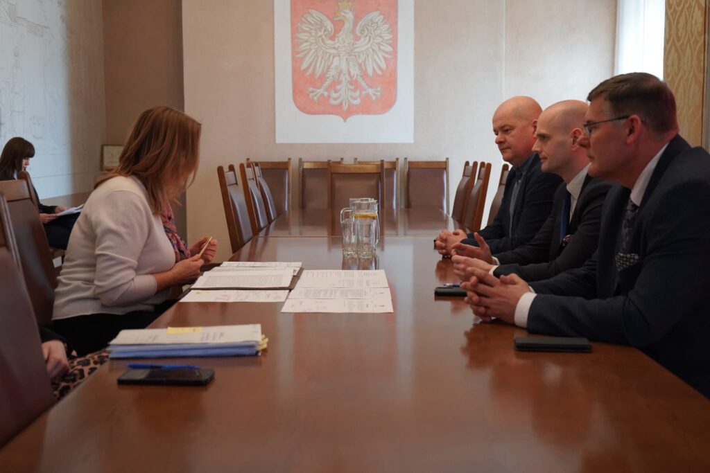 Grupa osób siedzących przy długim stole w sali konferencyjnej za Starostwem Powiatowym w Płocku, po lewej kobieta podpisująca dokumenty, po prawej trzej mężczyźni w garniturach, na ścianie projekt godła Polski.