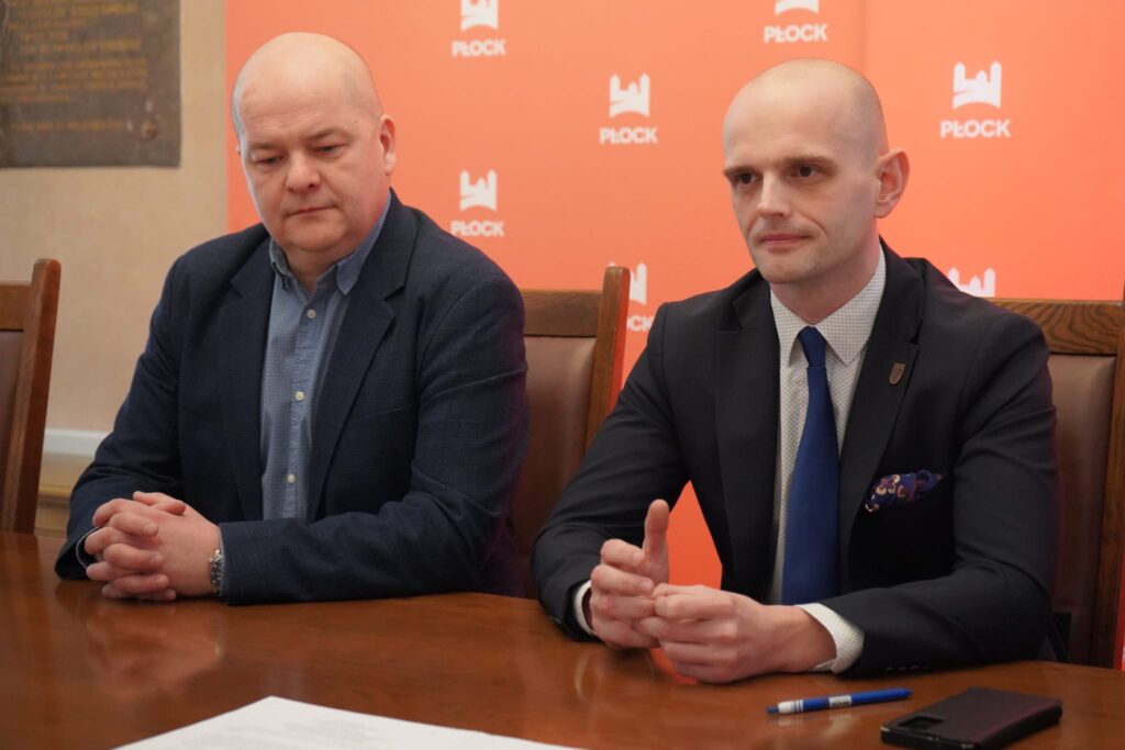 Dwóch mężczyzn w garniturach siedzi przy stole podczas podpisywania dokumentów, na tle pomarańczowego baneru z logo Płocka, w kontekście powstającego schronu z salą konferencyjną i Powiatowego Centrum Bezpieczeństwa za Starostwem Powiatowym w Płocku.