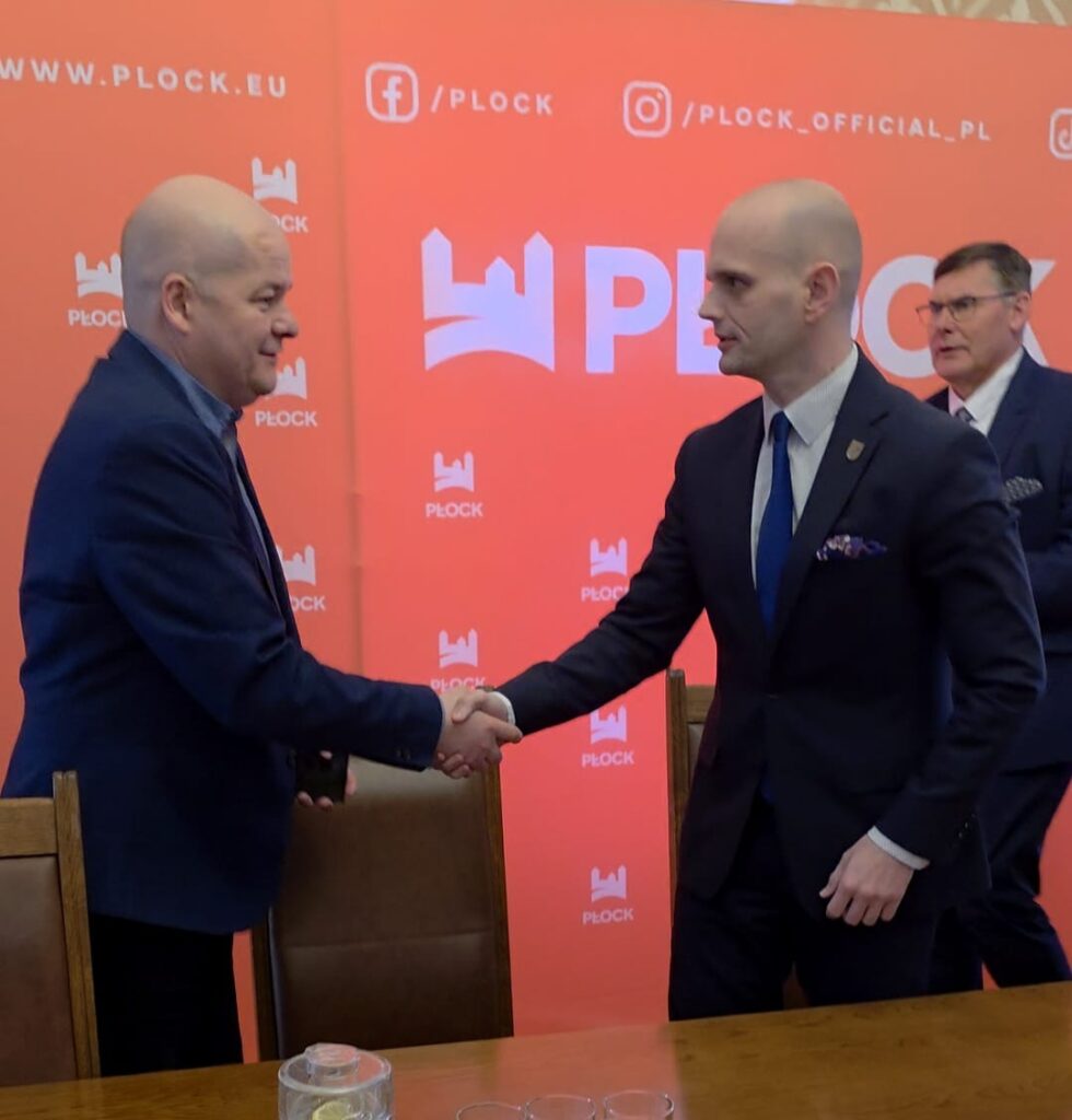 Dwóch mężczyzn w garniturach ściska sobie dłonie podczas spotkania, na tle pomarańczowego baneru z logo Płocka, zapowiadającego budowę schronu z salą konferencyjną i Powiatowego Centrum Bezpieczeństwa za Starostwem Powiatowym w Płocku.