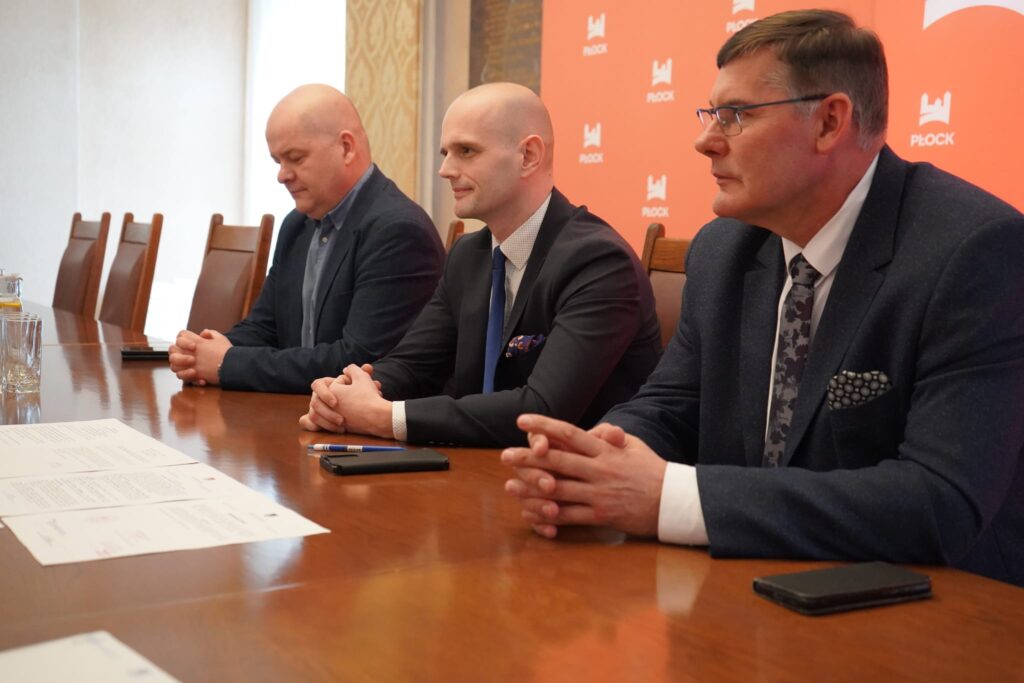 Trzech mężczyzn w garniturach siedzi przy długim stole w sali konferencyjnej podczas podpisywania dokumentów; w tle widoczny jest pomarańczowy baner z logo Płock.