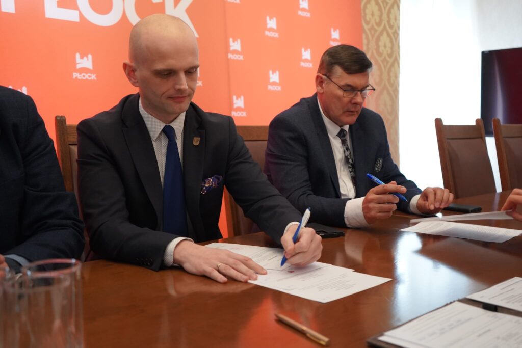 Dwóch mężczyzn w garniturach podpisuje dokumenty przy stole podczas oficjalnego spotkania, na tle pomarańczowego baneru z logo Płocka, w kontekście planowanego schronu z salą konferencyjną i Powiatowego Centrum Bezpieczeństwa w Płocku.