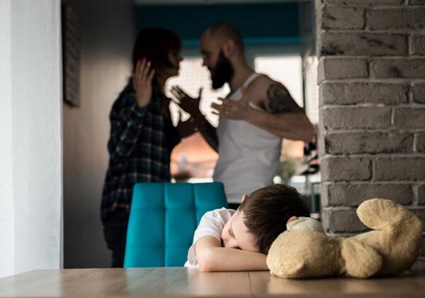 Dziecko siedzi przy stole z pluszowym misiem, a w tle dwie dorosłe osoby kłócą się i gestykulują.