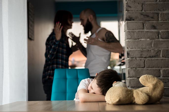 Dziecko siedzi przy stole z pluszowym misiem, a w tle dwie dorosłe osoby kłócą się i gestykulują.