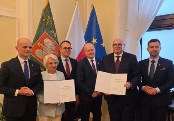 Grupa urzędników w garniturach stoi przy flagach Polski, Unii Europejskiej i lokalnej flagi, trzymając podpisane dokumenty dotyczące rozbudowy Wyszogrodzkiej.