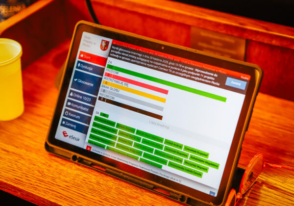 Tablet z interfejsem do głosowania i widocznymi wynikami (kolorowymi paskami) podczas XXI sesji Rady Powiatu Płockiego w kadencji 2024–2029, leżący na drewnianym stole obok żółtego kubka.